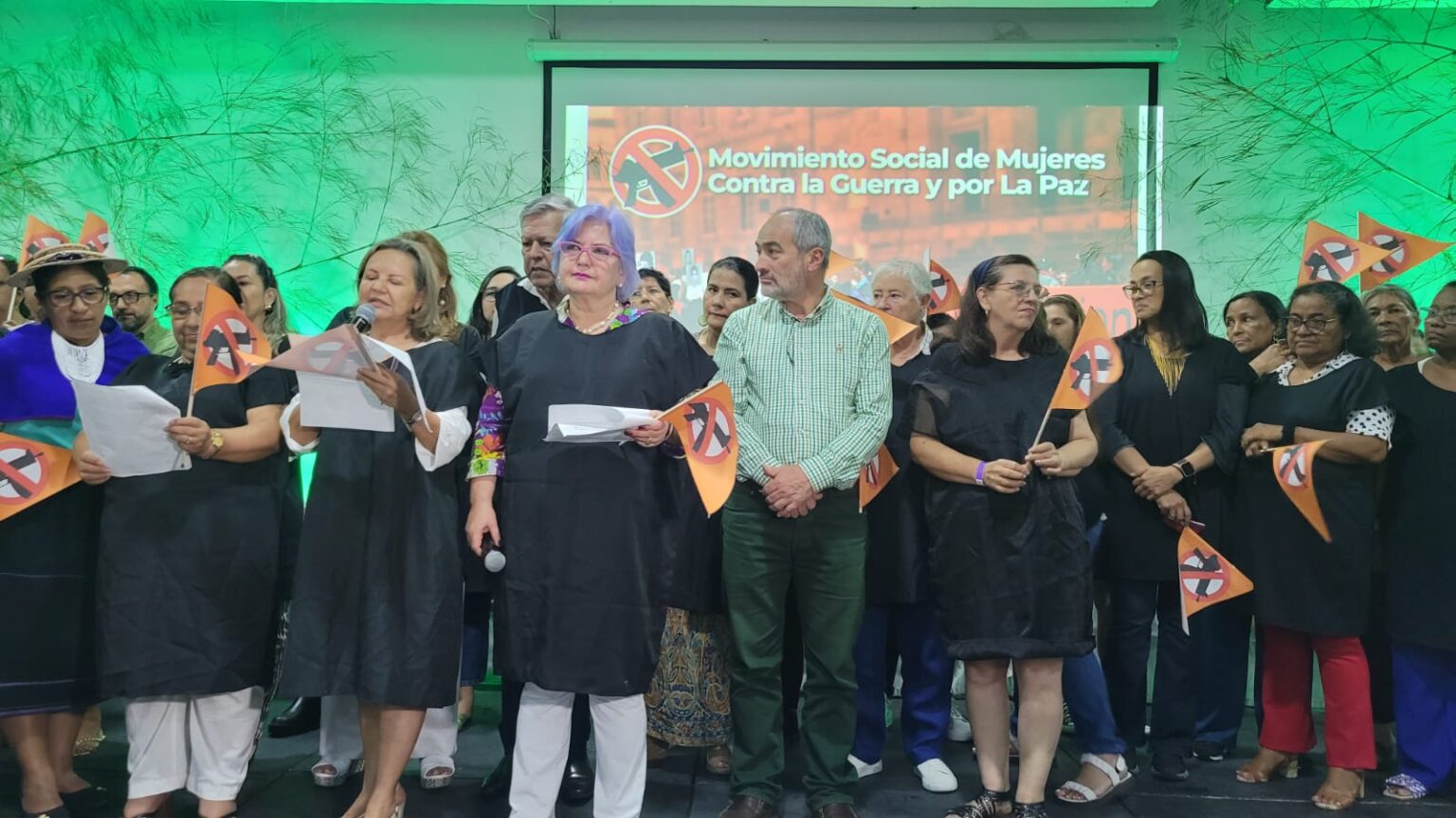 Mobilizing rally of the social women’s organization against war and for peace; a group of women and men on a stage with triangle shaped flags showing a machine gun in a red forbidden sign