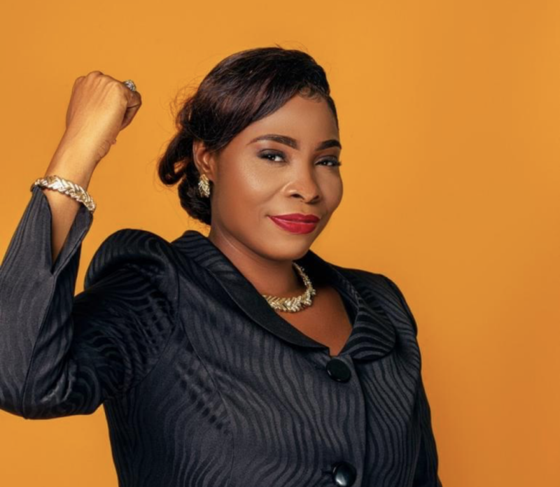 Jesula Blanc in a black pinstripe suit visible from the waist up in front of an orange photo studio background; African woman with a great confident smile and a fist up in the air