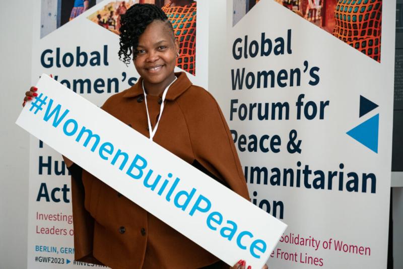 Vimbai Kapurura, African woman with a confident smile in a brown coat, standing in front of banners at the Global Woman’s Forum for Peace and Humanitarian Action in Berlin, holding a sign that reads #WomenBuildPeace